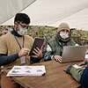 social workers masks sitting table using devices while talking black migrant tent